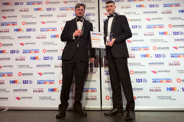 A photo at an awards ceremony of two men dressed in black suits standing side by side in front of a wall covered in company logos. The men are holding award plaques and statuettes.