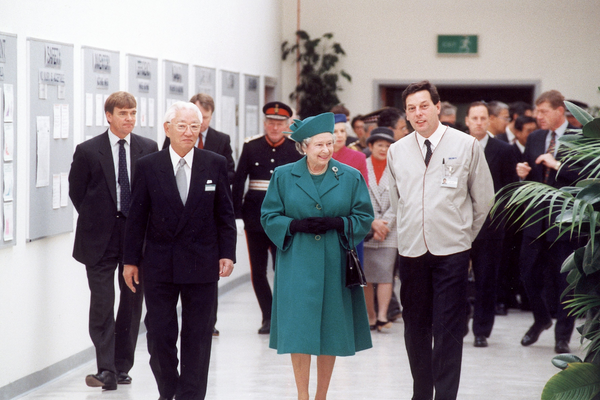 A photo from the opening of the Sony UK Technology centre building in 1992. At the centre of the photo is Her Late Majesty Queen Elizabeth the second dressed in a green dress wearing a matching hat who is walking down a white corrior. Next to her, to the left, walks one of Sony's founding fathers, Mr. Akio Morita dressed in a black suit. On Her Late Majesty's right is a local manager wearing a Sony uniform. There is a group of people walking behind them.