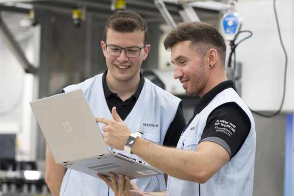 Two men stood side by side inside a factory. They are wearing Sony uniforms and looking at a computer screen together. On the sleeves of one man's shirt is the text 'A new way of working, engineering graduate'.