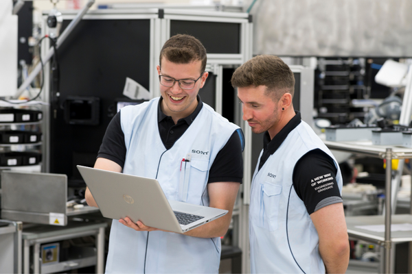 Two men wearing Sony uniforms are standing side by side inside a factory. They are looking at a computer screen together. On the sleeves of one man's shirt is the text 'A new way of working, engineering graduate'.