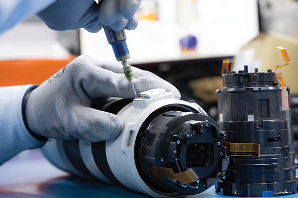 A close up photo of a camera lens repair process. In the frame, there are the hands of a technician wearing grey gloves and using a small blue screwdriver to work on a large white lens.