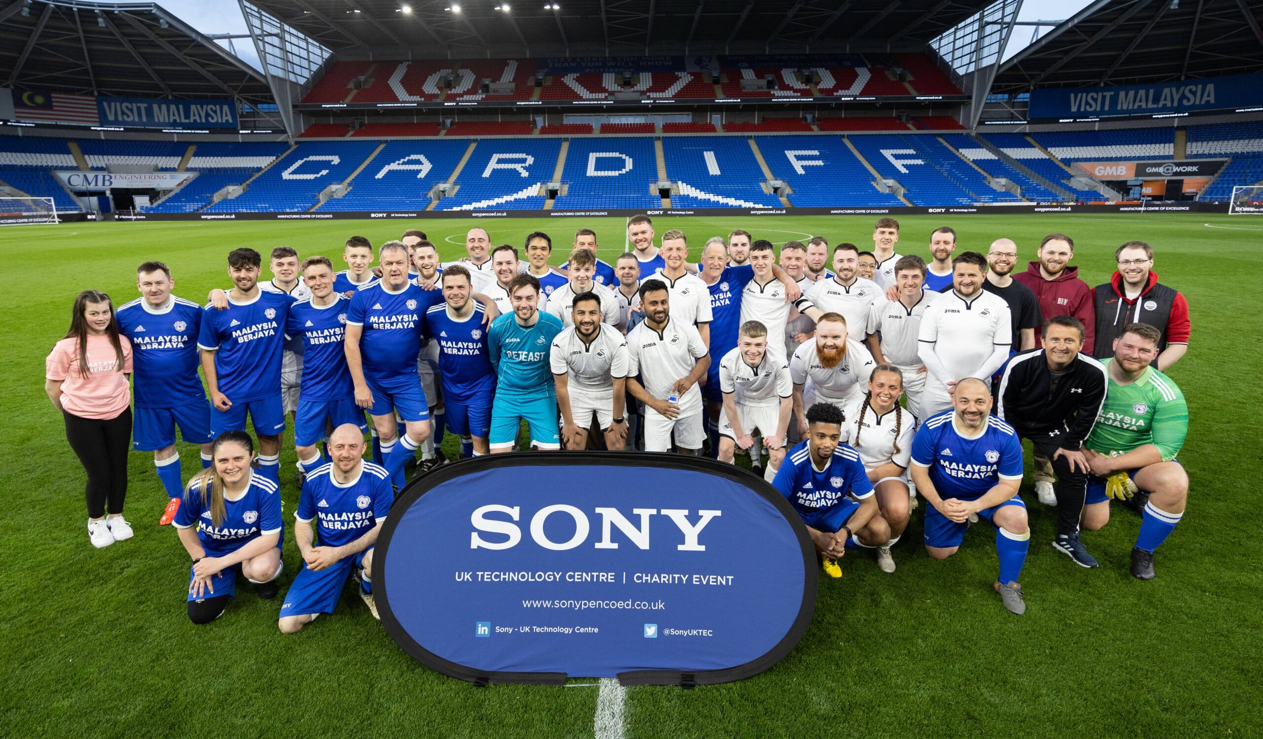 Two football teams dressed in white and blue uniforms standing on the Cardiff City Football Club pitch. In front them is a blue banner with the text 'Sony UK Technology Centre Charity Event'