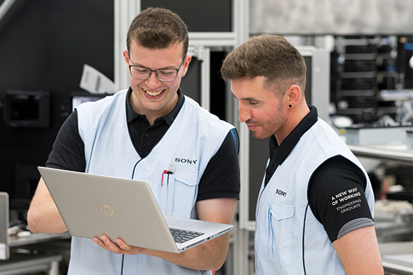 A photo of two men wearing Sony uniforms. One of them is holding an open laptop, and both men are looking at the screen. Behind them is the blurred outline of a manufacturing environment.