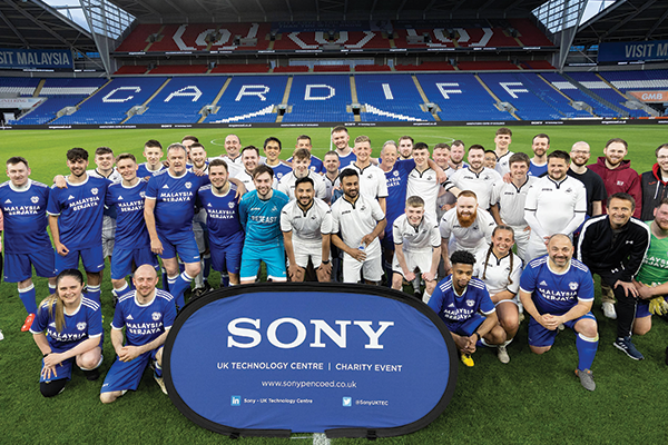 Two football teams dressed in white and blue uniforms standing on the Cardiff City Football Club pitch. In front them is a blue banner with the text 'Sony UK Technology Centre Charity Event'