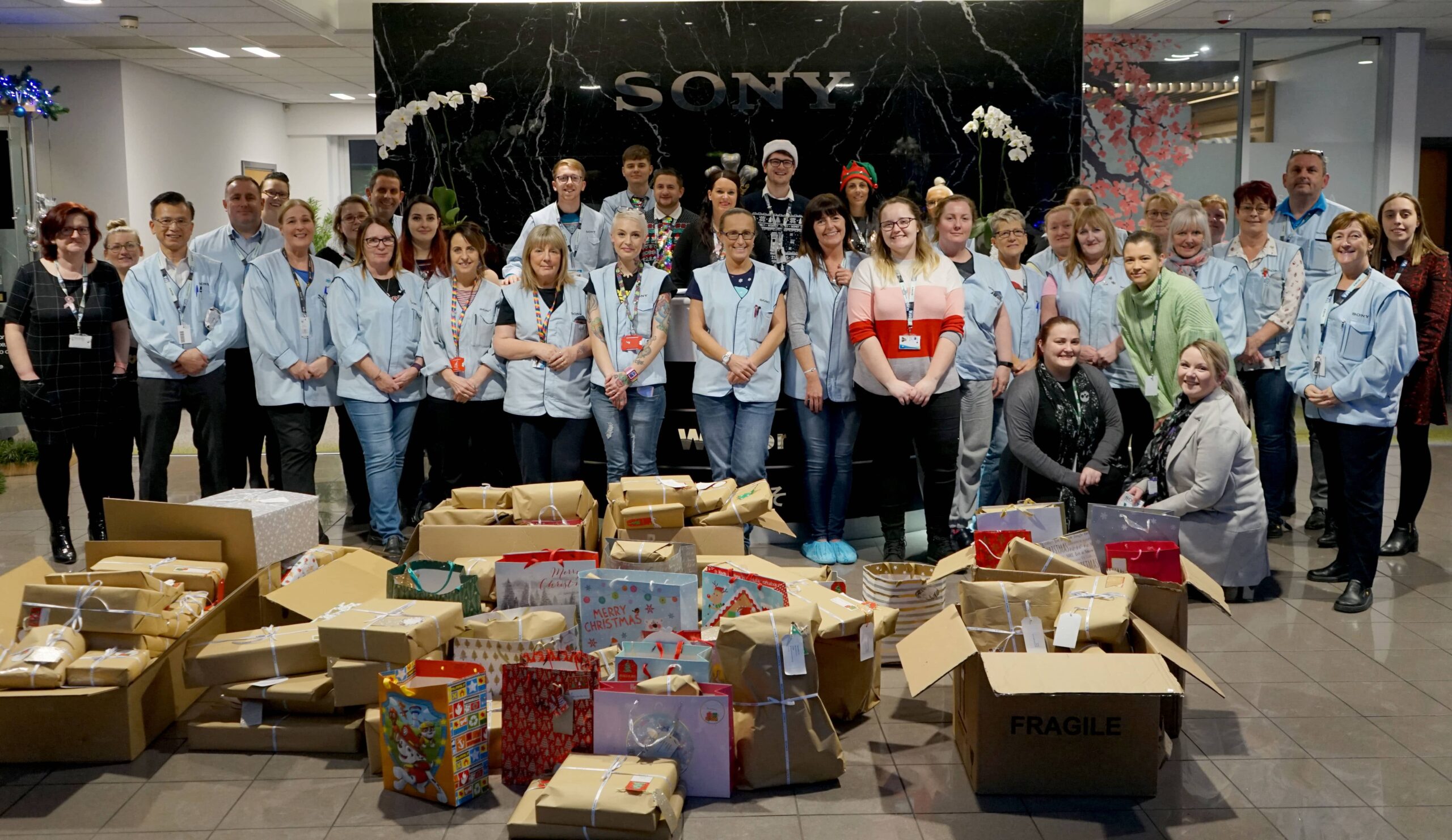 A large group of Sony employees standing in Sony UK TEC's reception. In front of them is a large pile of presents wrapped in brown paper.