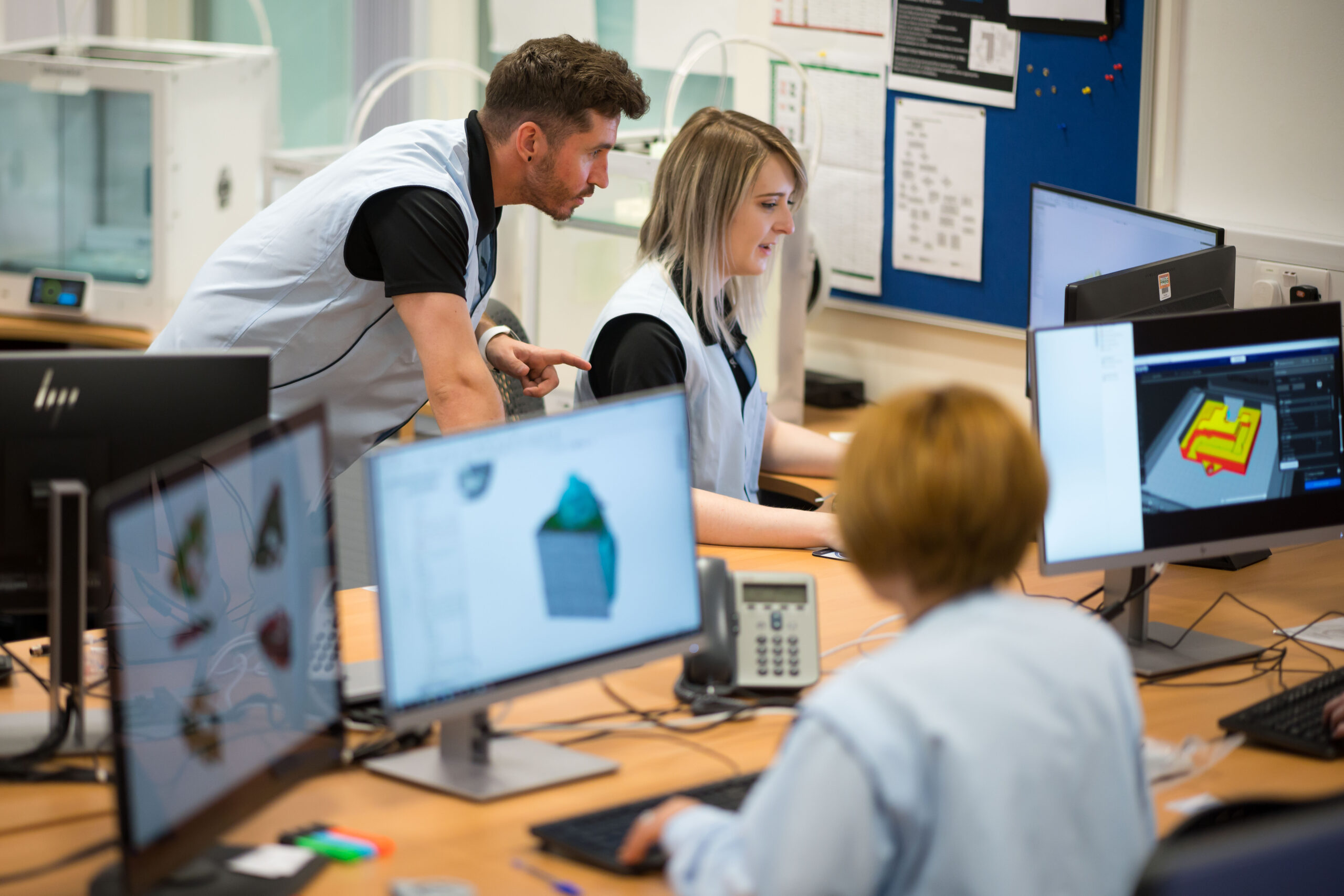 A photo of an office environment. There are two women and one man having a conversation. Various screens showing design software are next to them. Behind them, there are three white 3-D printers.