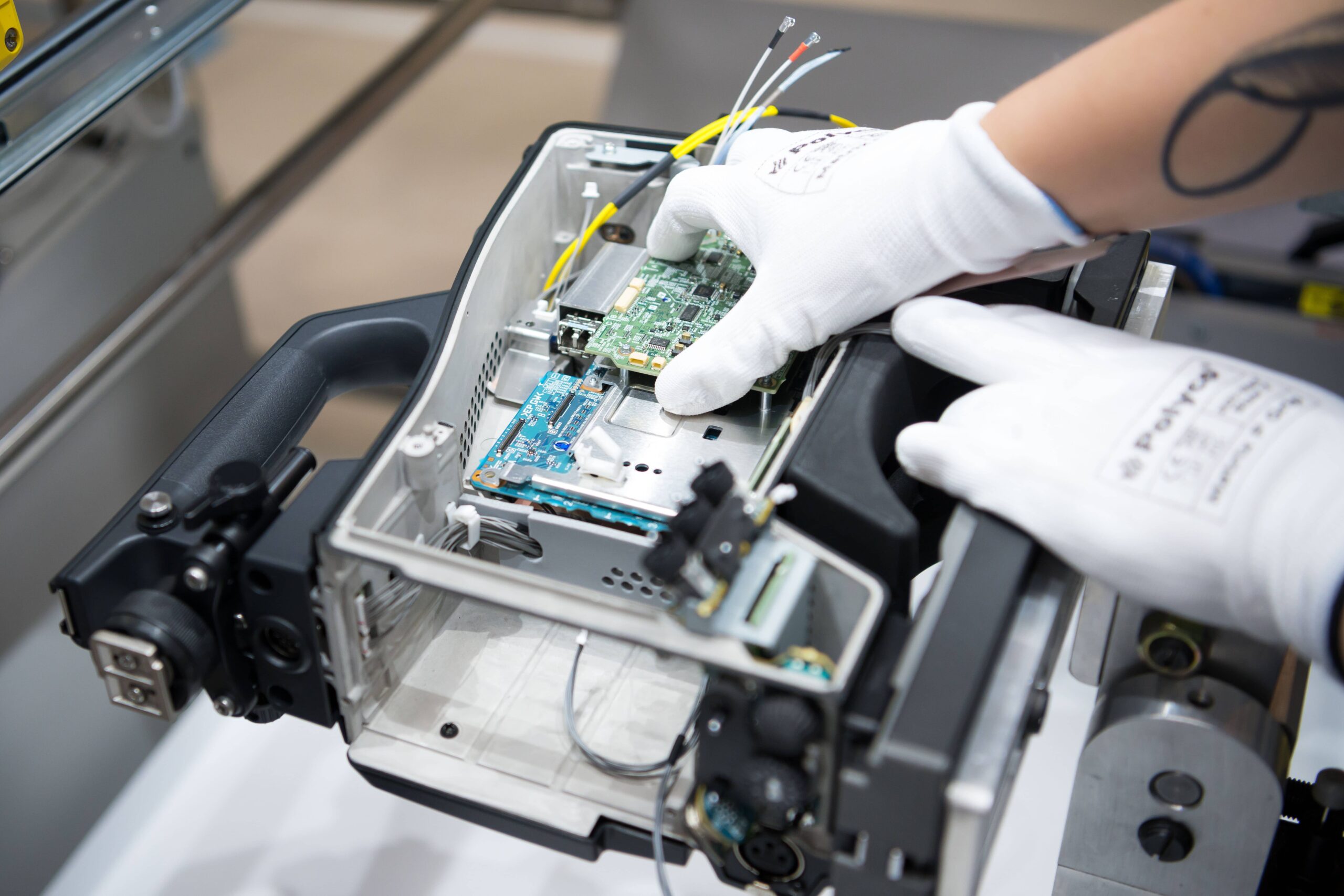 A close up photo of the inside of a Sony broadcast camera, showing the build process. There are two hands in the frame wearing white gloves, which are placing a circuit board inside the camera.