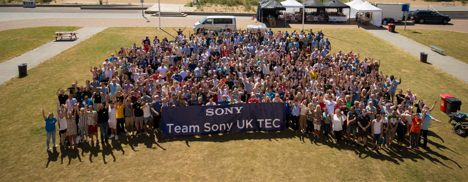 A photo of approximately 600 people standing in one group on a grassed area. In front of them is a blue banner with the text 