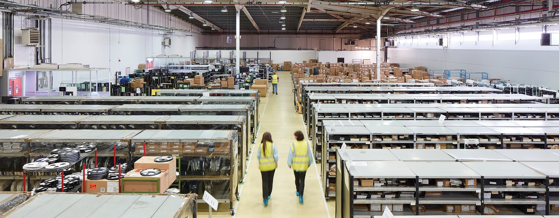 An areal photo of a warehouse. In the middle, there is a yellow walkway with three people. On the left and right of them are storage racks lined in several rows. At the background there are various brown boxes.