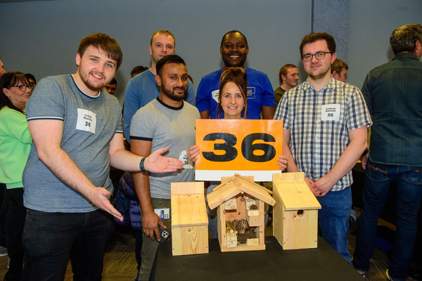 A photo of five men of various ethnicities and a woman standing together behind a table. On the table are three wooden units - a bird box, a bug hotel and a bat box.