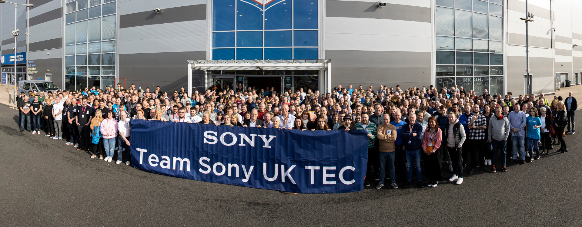 A photo of approximately 500 people standing in one group in front of the Cardiff City Football Club Stadium building. In front of them is a blue banner with the text 