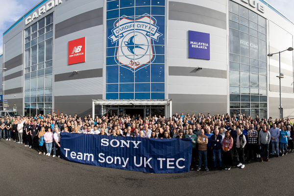 A photo of approximately 500 people standing in one group in front of the Cardiff City Football Club Stadium building. In front of them is a blue banner with the text 