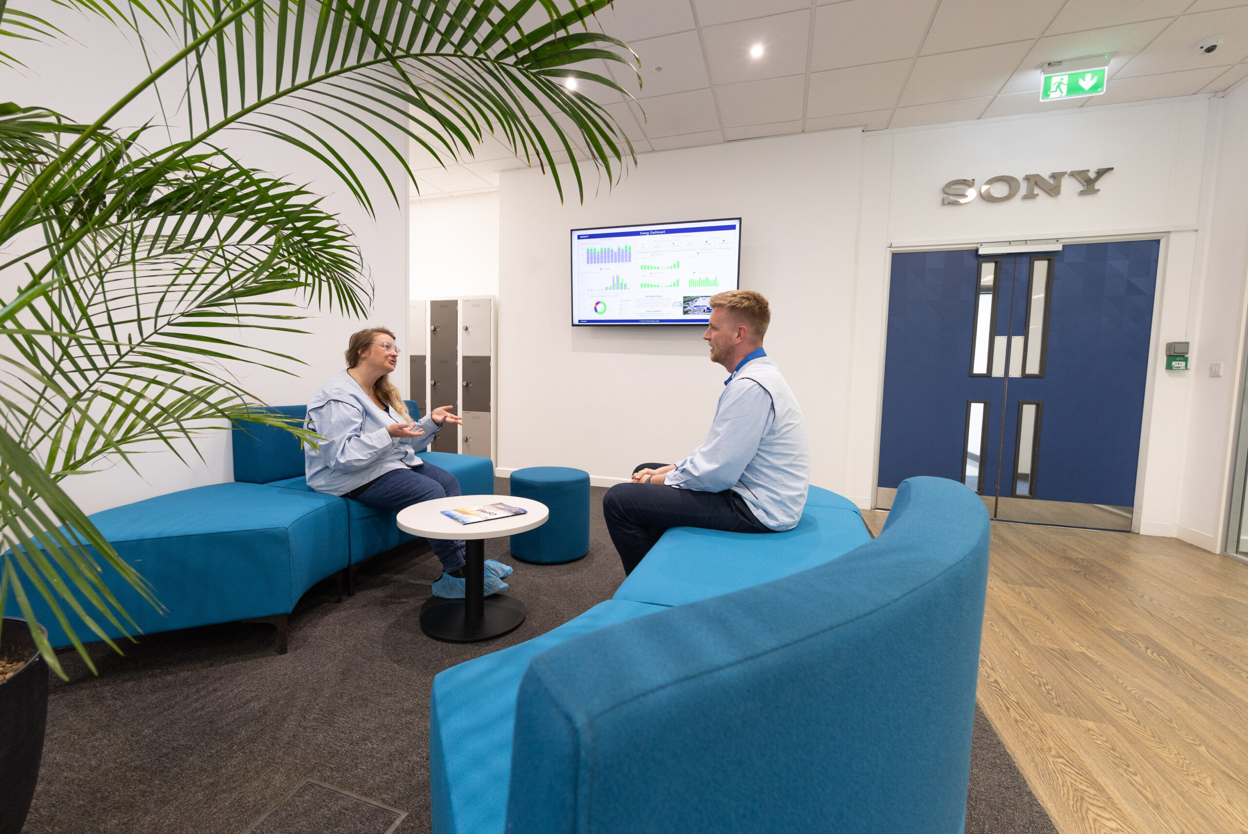 A man and a woman sitting on blue couches in a modern office, having a discussion. There is a screen with charts and data next to them.