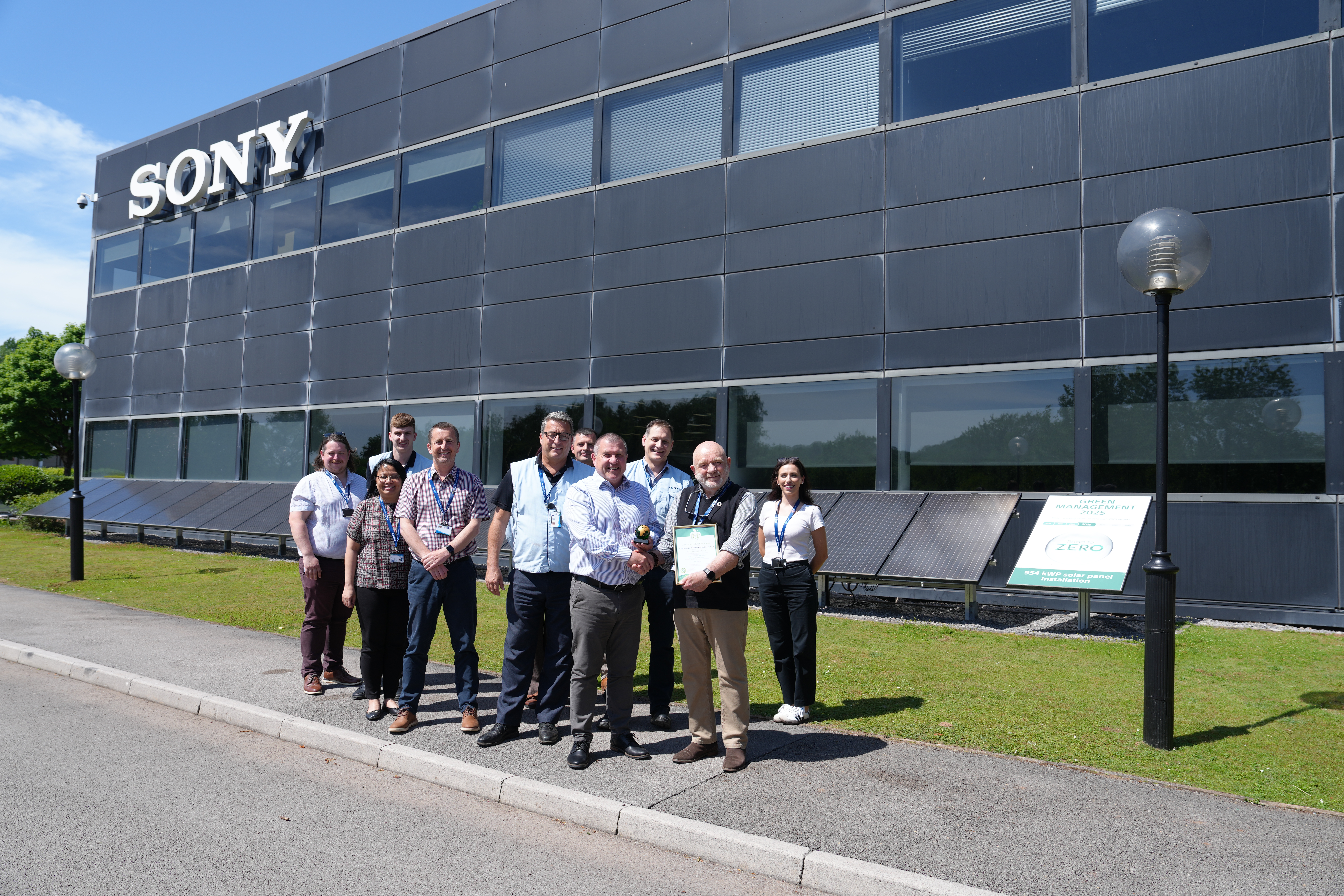 A group of people standing in front of a black building with a Sony logo on it's front. The people are smiling and holding a trophy and a framed certificate.